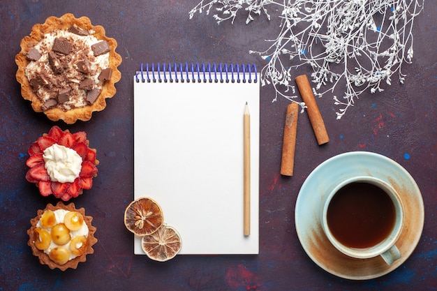 Top view of delicious cakes with cream chocolate and fruits with tea notepad on the dark surface