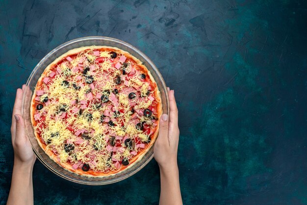 Вид сверху вкусная сырная пицца с оливками, сосисками в томатном соусе внутри стеклянной сковороды на голубом фоне.