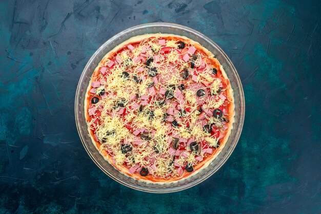 Вид сверху вкусной сырной пиццы с оливками и сосисками в томатном соусе внутри стеклянной сковороды на голубом столе.