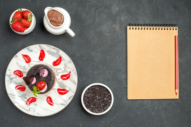 Top view delicious cheesecake with strawberry and chocolate on oval plate bowl of strawberries and chocolate a notebook on dark isolated background