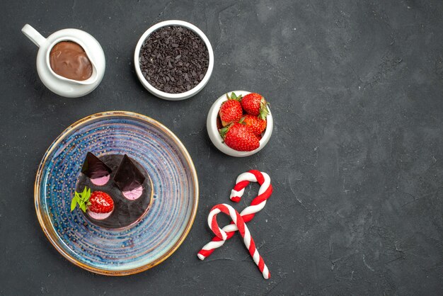 Top view delicious cheesecake with strawberry and chocolate on oval plate bowls with strawberries chocolate xmas candies on dark isolated background