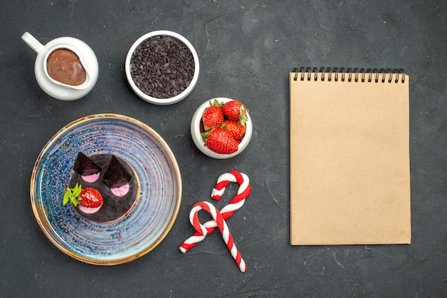 Top view delicious cheesecake with strawberry and chocolate on oval plate bowls with strawberries chocolate xmas candies a notebook on dark isolated background