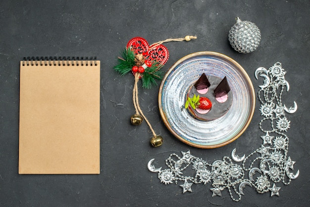 Vista dall'alto deliziosa cheesecake con fragole e cioccolato su un piatto d'argento ovale ornamenti di natale un taccuino su sfondo scuro isolato