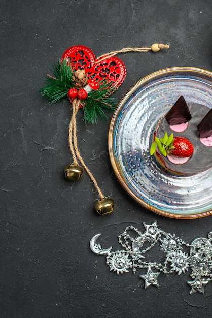 Top view delicious cheesecake with strawberry and chocolate on oval silver plate xmas pendants on dark isolated background