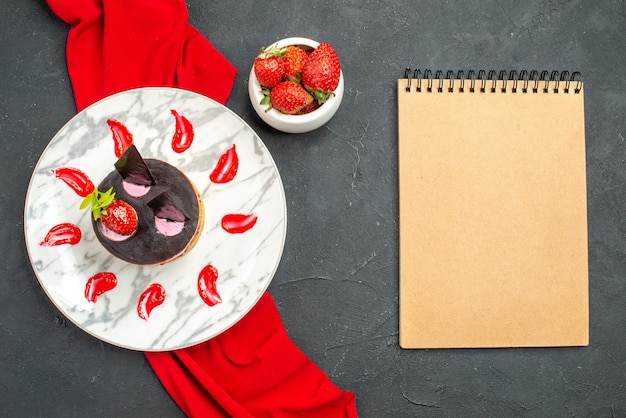 Top view delicious cheesecake with strawberry and chocolate on plate red shawl bowl with strawberries a notebook on dark isolated background