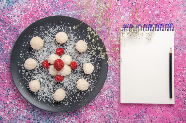 Top view of delicious coconut candies sweet balls with notepad on pink surface