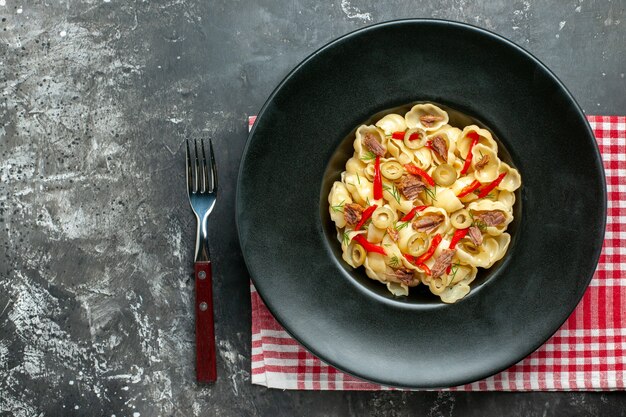 Вид сверху вкусной конкильи с овощами и зеленью на тарелке и ножом на красном полосатом полотенце на сером фоне