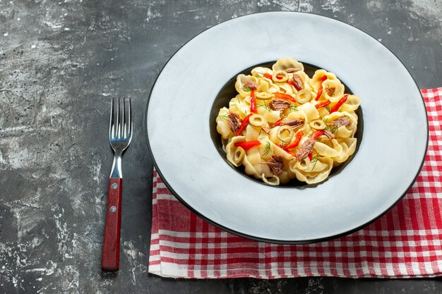 Top view of delicious conchiglie with vegetables and greens on a plate and knife on red stripped towel on the left side on gray background