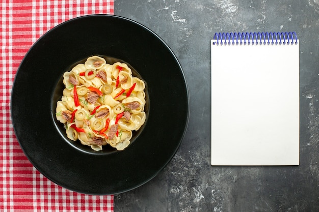 Top view of delicious conchiglie with vegetables and greens on a plate and knife on red stripped towel next to notebook on gray background