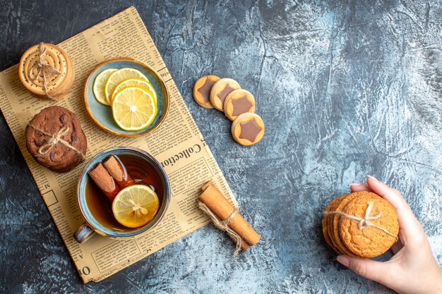 Top view of delicious cookies and hand holding a cup of black tea with cinnamon on an old newspaper on dark background
