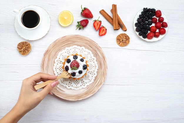 top view of delicious creamy cake with berries getting eat by female with cinnamon coffee on light desk,  cake sweet