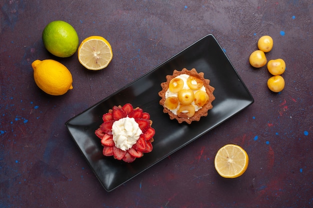 Вид сверху вкусных сливочных пирожных внутри тарелки со свежими лимонами на темной поверхности