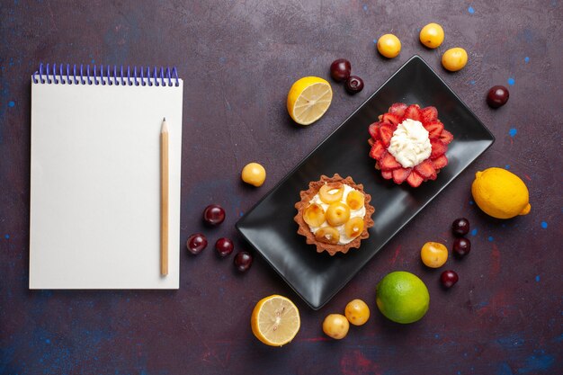 Top view of delicious creamy cakes inside plate with fresh lemons notepad and fruits on dark surface