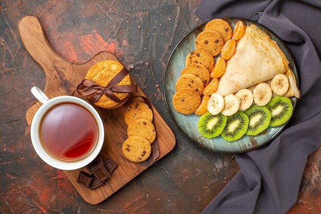 Top view of delicious crepe chopped citrus fruits cookies on dark towel and chocolate bars a cup of black tea on mixed color