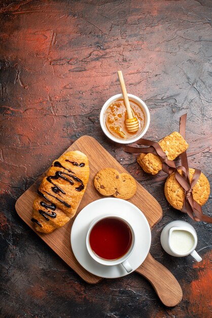 Top view of delicious croisasant a cup of black tea on a wooden cutting board honey stacked cookies milk on a dark surface