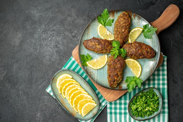 Top view of delicious cutlets served with lemon and green on wooden cutting board on yellow stripped towel on black background