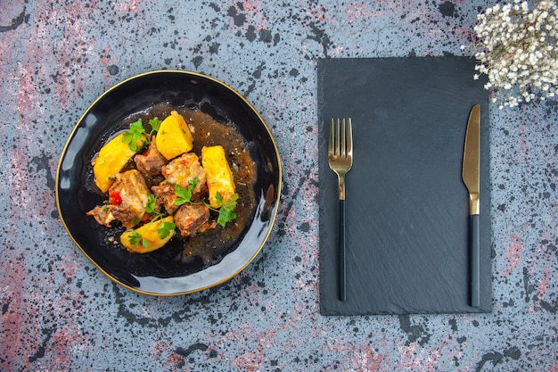 Top view of delicious dinner with meat potatoes served with green in a black plate and cutlery set on cutting board flower on mix colors background