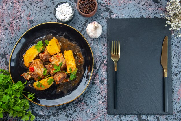 Top view of delicious dinner with meat potatoes served with green in a black plate and cutlery set on cutting board flower spices garlic on mix colors background