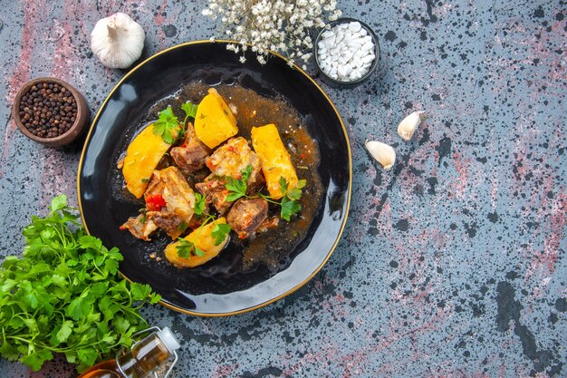 Top view of delicious dinner with meat potatoes served with green in a black plate and garlic spices fallen oil bottle on mix colors background