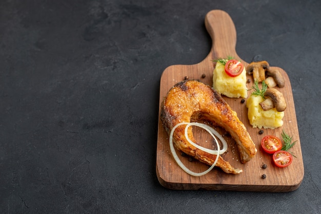 Top view of delicious fried fish meal and mushrooms tomatoes greens on wooden cutting board on the left side on black surface