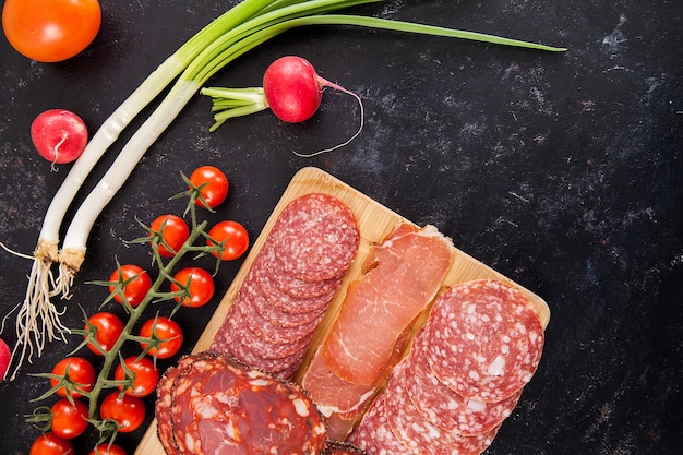 Вид сверху на вкусные здоровые мясные закуски на деревянной доске рядом с помидорами черри, редисом и зеленым луком