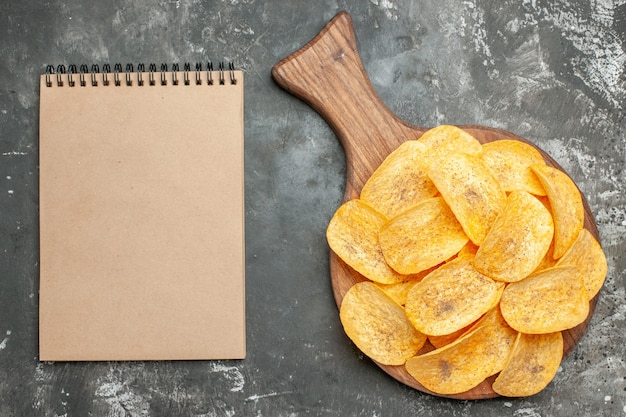 Top view of delicious homemade chips on wooden cutting board and notebook on gray background