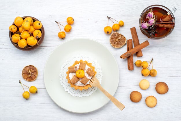 Top view of delicious little cake along with yellow cherries cinnamon coookies and tea on light, cookie cake biscuit sweet