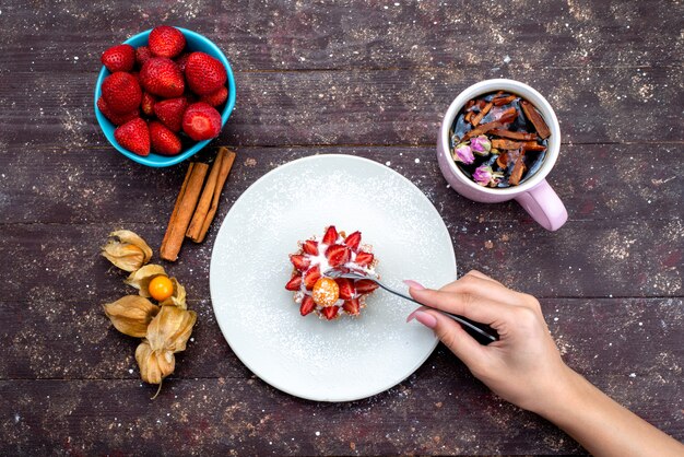 A top view delicious little cake with cream and fresh sliced fruits along with cinnamon and strawberries on the brown desk fruit cake biscuit