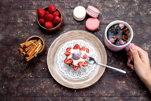 A top view delicious little cake with cream and fresh sliced fruits along with cinnamon tea drinking by female and macarons on the brown desk fruit cake