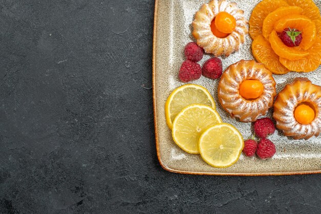 Top view delicious little cakes with lemon slices and tangerines on the dark background fruit biscuit sweet tea cookie