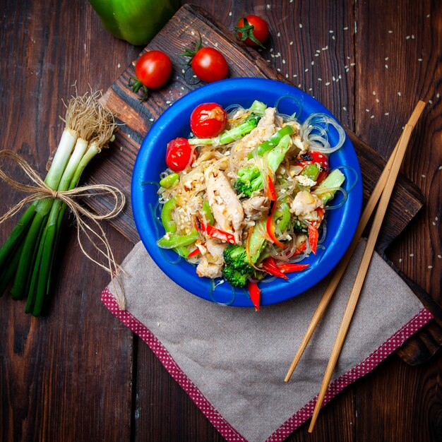 Top view delicious meal in blue plate with green onions, tomatoes and chopsticks on dark wooden background.