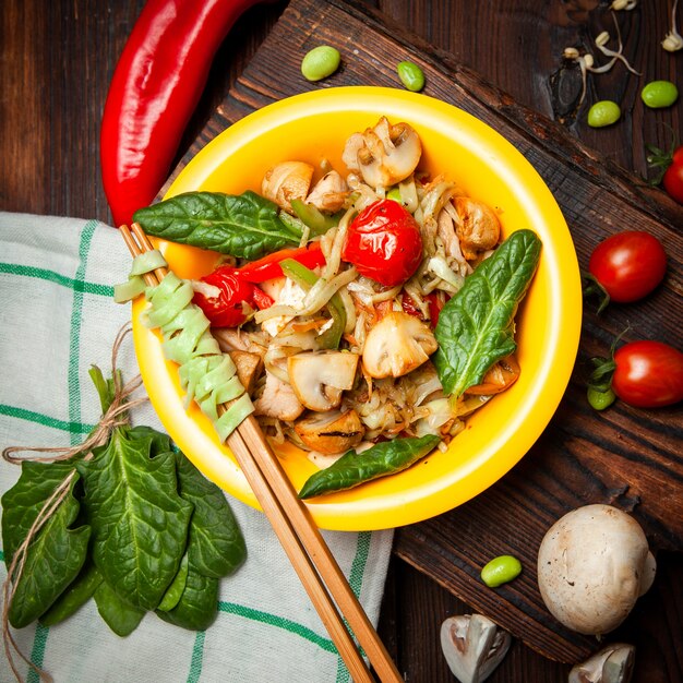 Top view delicious meal in yellow plate with red pepper, tomatoes, chopsticks on wood, cloth and dark wooden background.