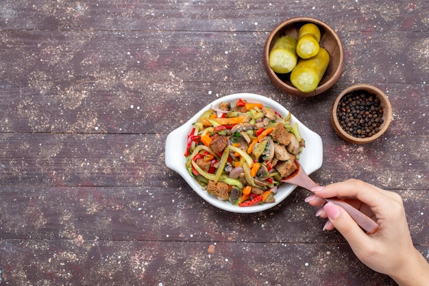 Вид сверху вкусного мясного салата с нарезанным мясом и приготовленными овощами вместе с солеными огурцами на коричневом столе, мясное блюдо для еды