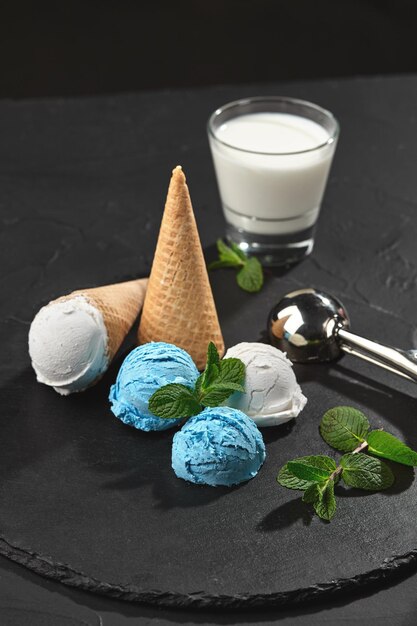 Top view of a delicious natural creamy and blueberry ice cream decorated with mint, and classic waffle cones are served on a stone slate, standing on a dark table over a black background. Metal scoop