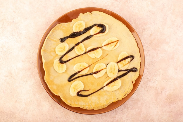 A top view delicious pancakes designed with chocolate and bananas inside round plate on the pink desk food meal dessert pastry