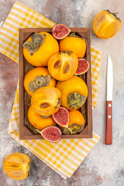 Top view delicious persimmons and cut figs in wood box yellow kitchen towel a knife on nude background