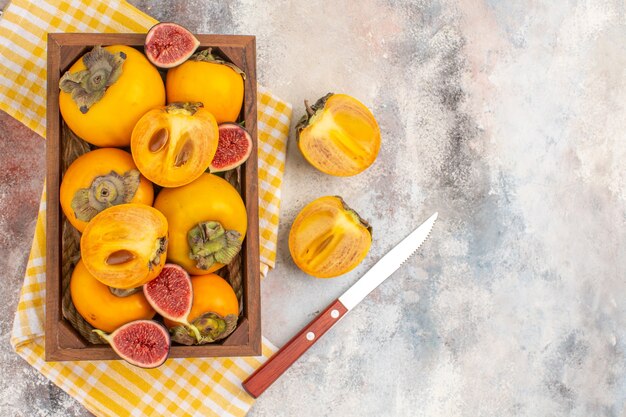 Top view delicious persimmons and cut figs in wood box yellow kitchen towel a knife on nude background