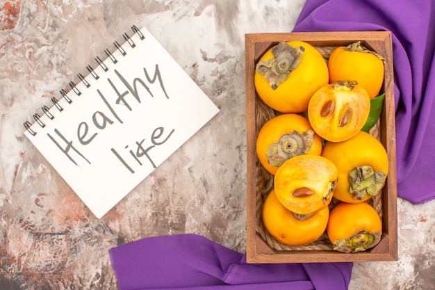 Top view delicious persimmons in a wood box healthy life written on notebook purple shawl on nude background