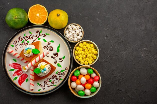 Top view delicious pie slices with candies and fresh tangerines on dark background cake sweet biscuit pie fruit