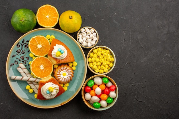 Top view delicious pie slices with candies and fresh tangerines on the dark background fruit cake sweet biscuit pie