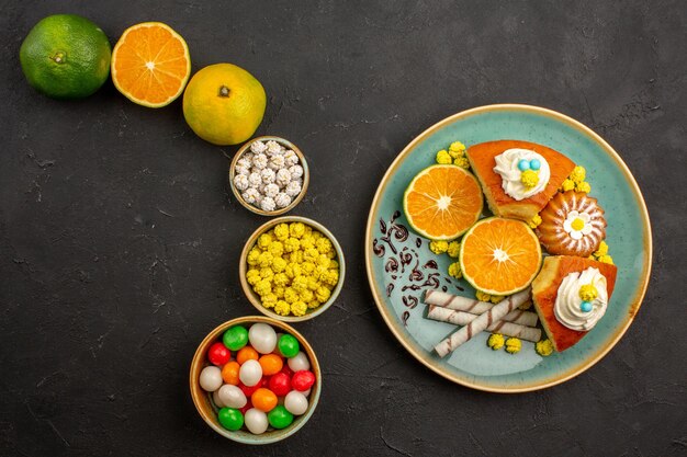 Top view delicious pie slices with candies and fresh tangerines on dark desk fruit cake sweet biscuit pie