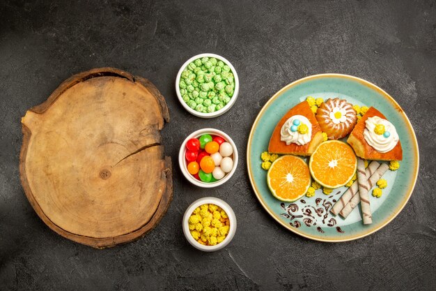 Top view delicious pie slices with tangerines and candies on a dark-grey background fruit candy cake pie dough tea