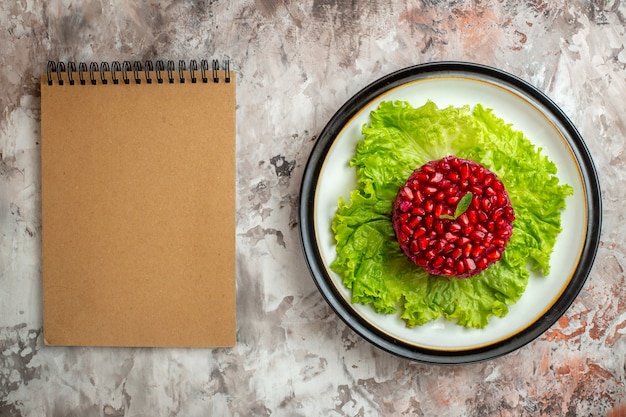 Vista dall'alto deliziosa insalata di melograno a forma rotonda su insalata verde con blocco note sullo sfondo chiaro Foto Gratuite