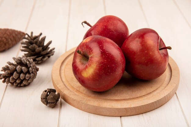 Top view of delicious red apples on a wooden kitchen board with pine cones isolated on a white wooden surface