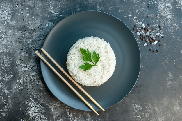 Вид сверху вкусной рисовой еды, подаваемой с зеленью и палочками для еды на черной тарелке с перцем на темном фоне