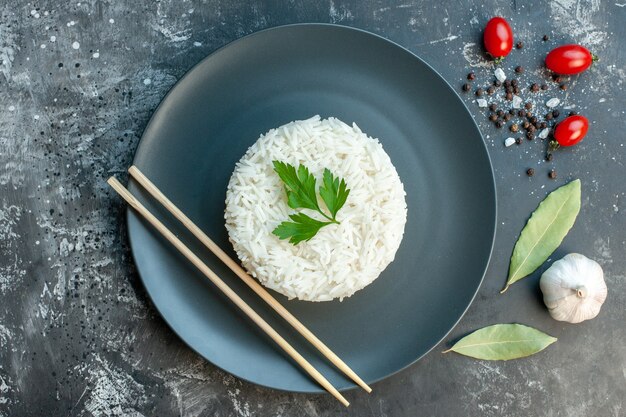 Вид сверху вкусной рисовой еды, подаваемой с зеленью и палочками для еды на черной тарелке, перец, чеснок, помидоры на темном фоне