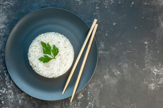 Вид сверху вкусной рисовой еды, подаваемой с зеленью и палочками для еды на черной тарелке с правой стороны на темном фоне