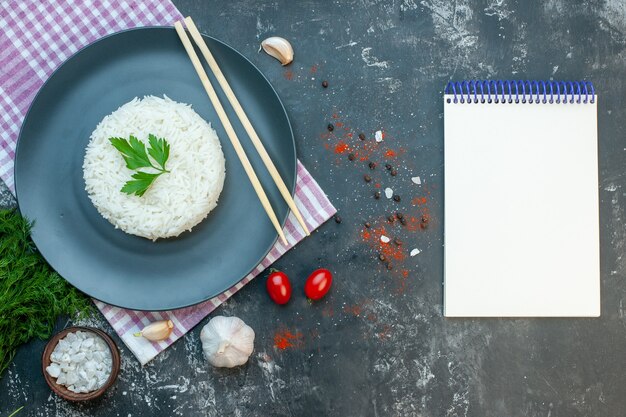 Вид сверху вкусной рисовой муки, подаваемой с зелеными деревянными чопстиками на черной тарелке на фиолетовом полосатом полотенце рядом с солеными помидорами с чесноком рядом с помидорами на черном фоне