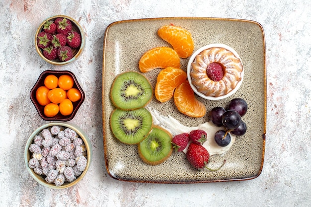 Вид сверху вкусные нарезанные фрукты с тортом и конфетами на белом фоне фруктовый свежий чай, конфета, торт, бисквит