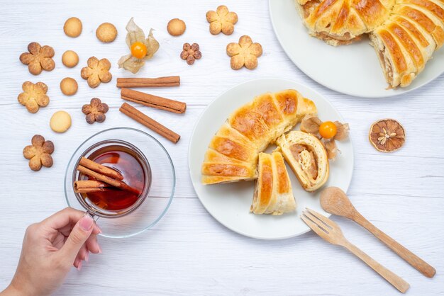 Top view of delicious sliced pastries inside plate with filling along with tea cinnamon and cookies on light, pastry cookie biscuit sweet